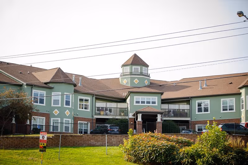 Exterior view of a two-story senior living facility building with green walls and brown roof. The building features a central tower-like structure with diamond-shaped decorations and multiple windows. There is a grassy area with bushes and a sign in front of the building, along with several parked cars visible behind a fence.