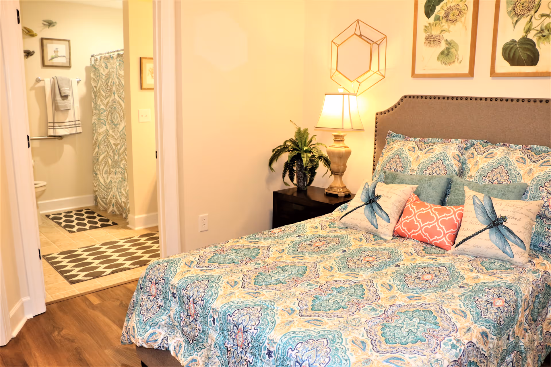 A cozy bedroom with a patterned bedspread in shades of blue, green, and beige. The bed has decorative pillows, including two with dragonfly designs and one coral-colored pillow. A nightstand beside the bed holds a lamp and a potted plant. On the wall above the bed are two framed botanical prints and a geometric wall decoration. An open door reveals a bathroom with a patterned shower curtain, towels hanging on a rack, and a tiled floor with two rugs.