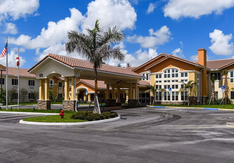 Exterior view of Elegance at Lake Worth senior living facility showing a large building with beige and orange walls, multiple windows, a covered entrance with stone pillars, palm trees, and a clear blue sky with scattered clouds.