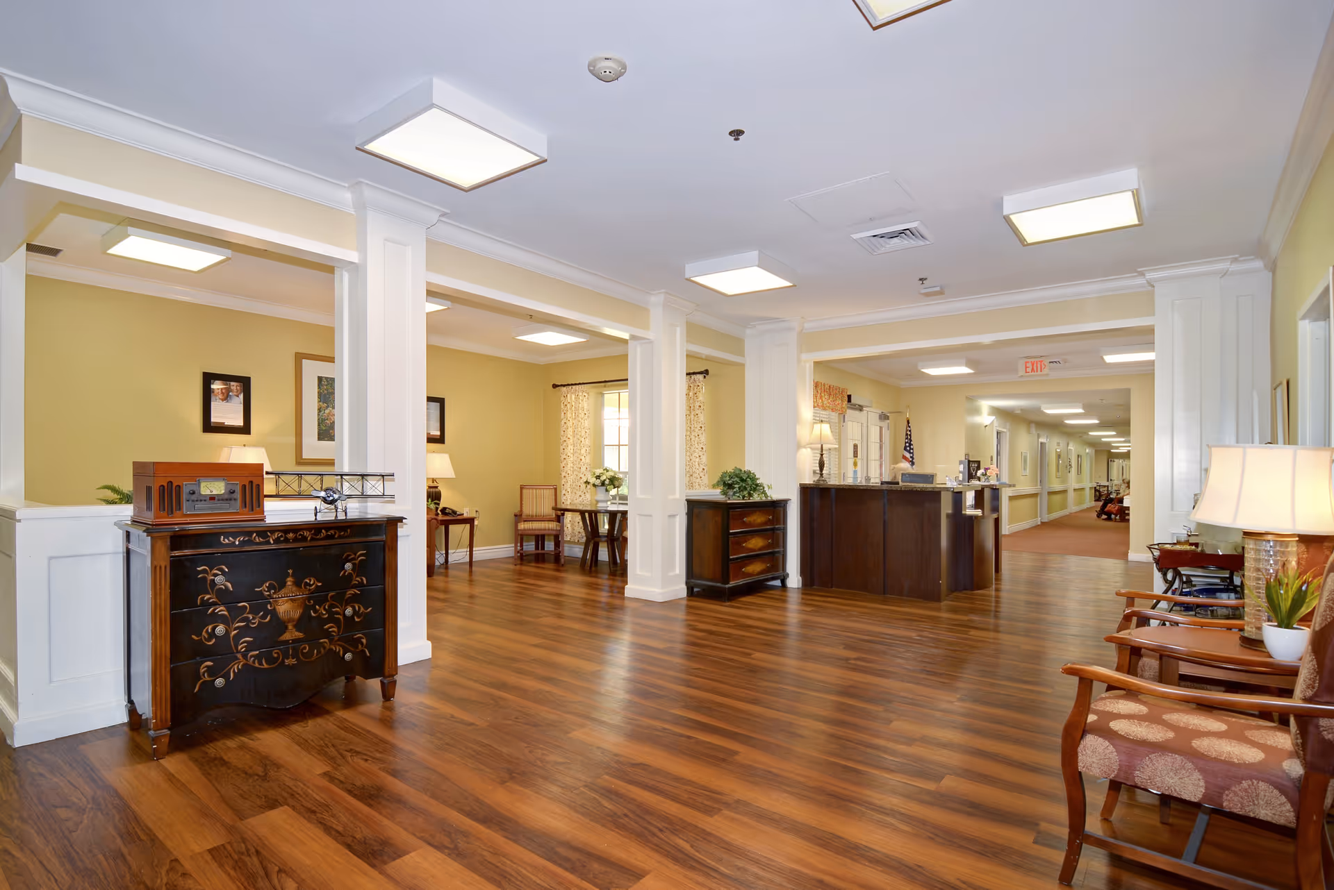 Interior view of a senior living facility lobby area with wooden flooring, cream-colored walls, decorative furniture including a chest with a vintage radio and model airplane, chairs with patterned upholstery, lamps, and a reception desk in the background. The space is well-lit with ceiling lights and has framed pictures on the walls.