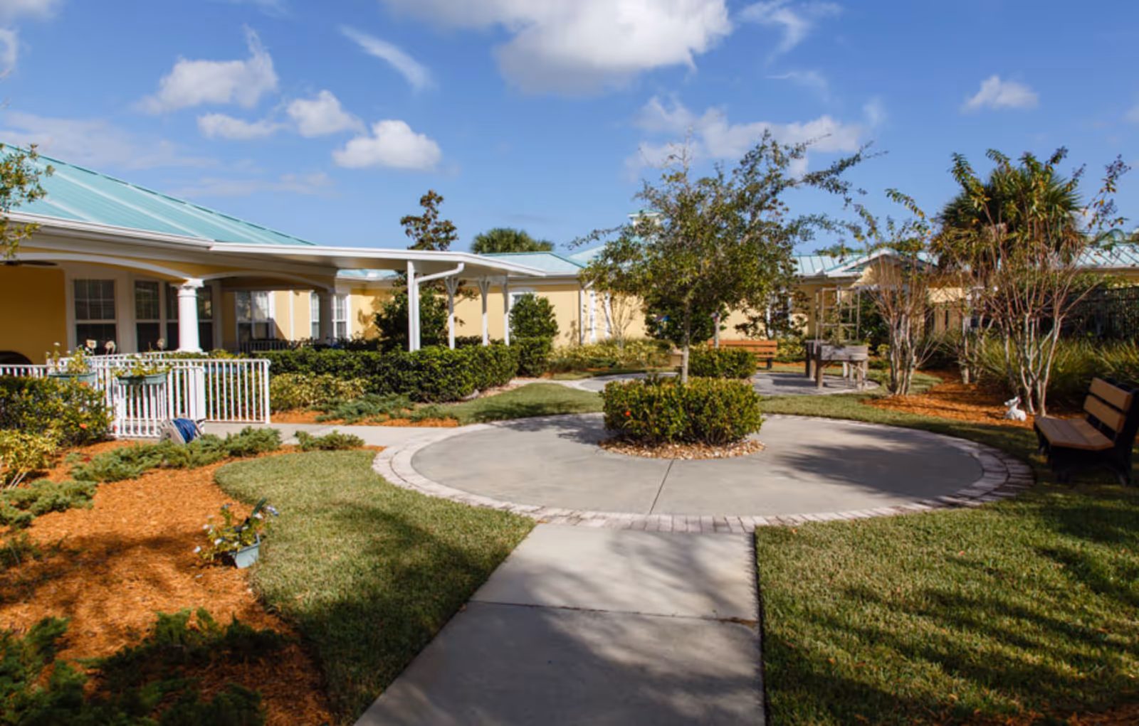 Outdoor courtyard area at The Brennity at Vero Beach Senior Living featuring a circular paved walkway with a small tree and bushes in the center, surrounded by green grass, landscaped mulch beds, benches, and a covered porch attached to a yellow building with a light blue roof under a partly cloudy sky.