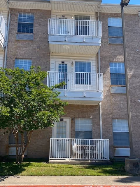 Exterior view of a three-story brick apartment building with white balconies on the second and third floors and a small fenced patio on the ground floor. There is a tree on the left side and a streetlamp on the right side.