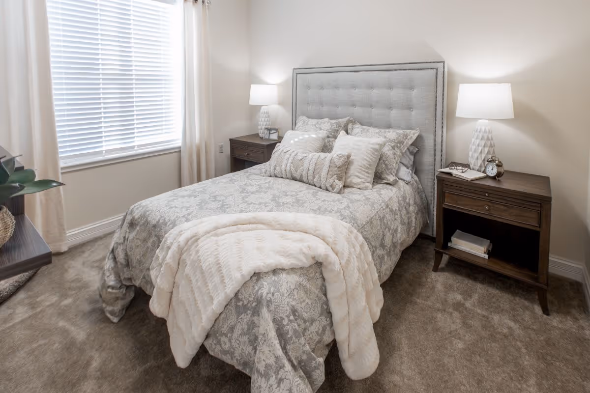 A cozy bedroom with a neatly made bed featuring a patterned gray and white bedspread, multiple decorative pillows, and a soft white throw blanket. The bed has a tufted gray headboard. On each side of the bed are dark wooden nightstands with white table lamps. A window with white blinds and light curtains is on the left wall, and the floor is carpeted in a neutral tone.