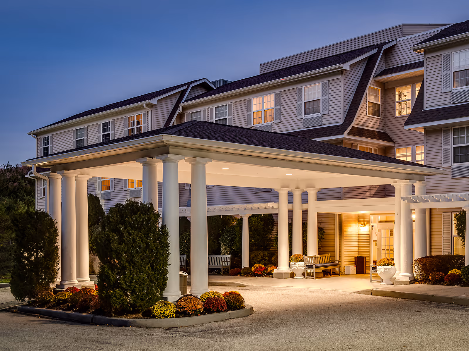 Exterior view of Atria Briarcliff Manor senior living facility at dusk, showing the entrance with a covered driveway supported by white columns, well-maintained landscaping with bushes and flowers, and warm lights glowing from the windows and entrance area.