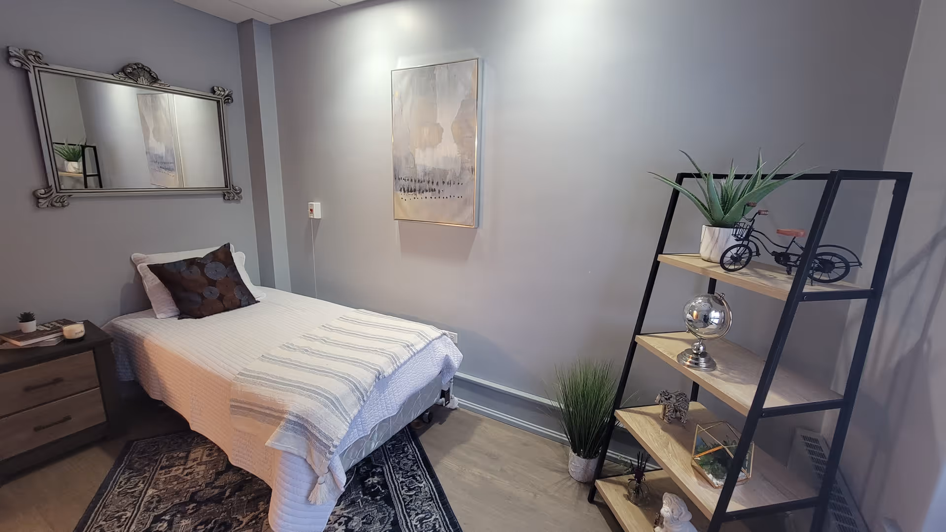 A neatly arranged bedroom with a single bed covered in a white quilt and a striped throw blanket. There is a dark pillow on the bed. To the left of the bed is a wooden nightstand with a small plant, a candle, and a book. Above the bed is a decorative mirror. On the wall opposite the bed hangs an abstract painting. To the right is a black metal and wood shelving unit with decorative items including a potted plant, a small bicycle model, and other ornaments. The room has light-colored walls and wood flooring with a patterned rug partially under the bed.