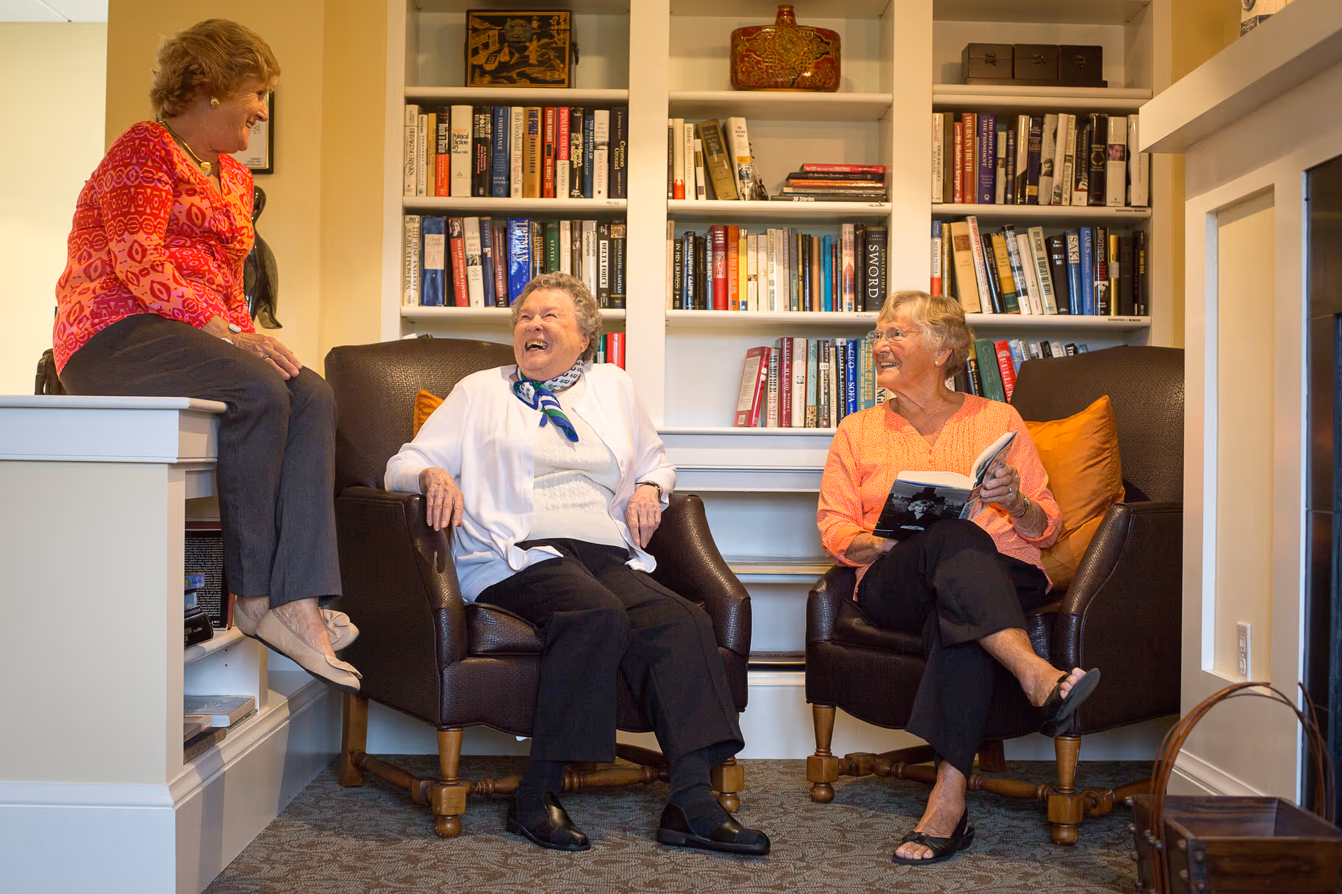 Three elderly women sitting and chatting in a cozy room with built-in bookshelves filled with books. Two women are seated in brown armchairs, one holding a book, while the third woman sits on a ledge beside them. They are smiling and appear to be enjoying each other's company.