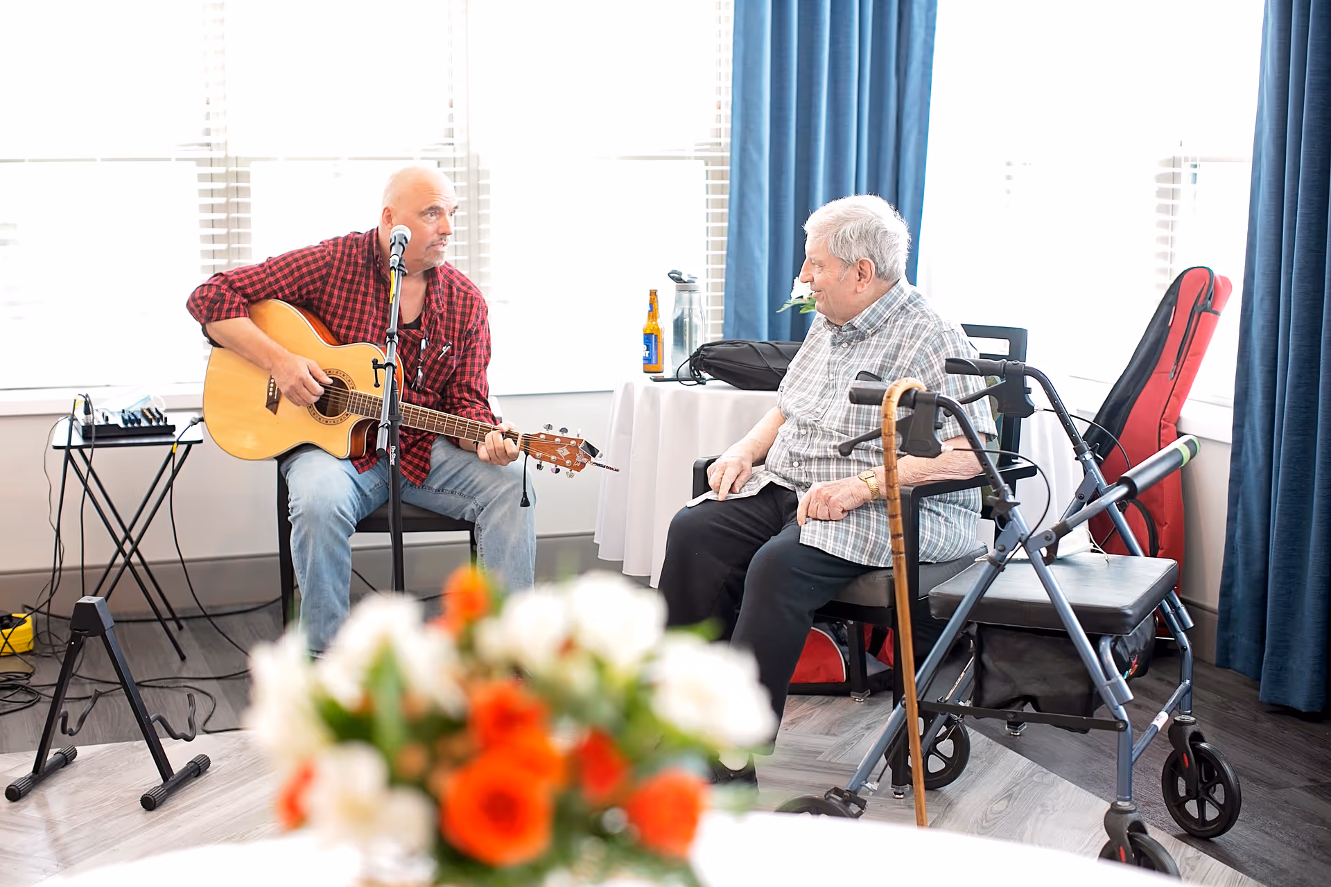An elderly man sitting in a chair with a walker nearby, listening to a man playing an acoustic guitar and singing into a microphone in a bright room with large windows and blue curtains.
