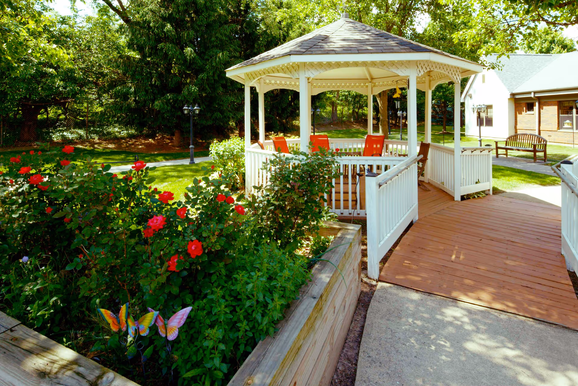 A bright outdoor garden area featuring a white wooden gazebo with a shingled roof and orange chairs inside. Surrounding the gazebo are lush green plants and red flowers, with a wooden raised garden bed in the foreground. A paved walkway leads up to the gazebo, and a wooden bench is visible near a brick building in the background under a sunny sky.