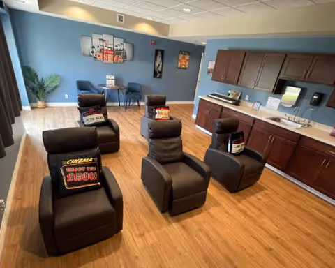 A cozy room with five black reclining chairs arranged in two rows facing a wall with cinema-themed decorations. Each chair has a pillow with the words 'Cinema Holiday The Show'. The room has wooden flooring, blue walls, and a kitchenette area with dark wooden cabinets, a sink, and a countertop on the right side. There is a small table with two chairs and a potted plant in the back corner.