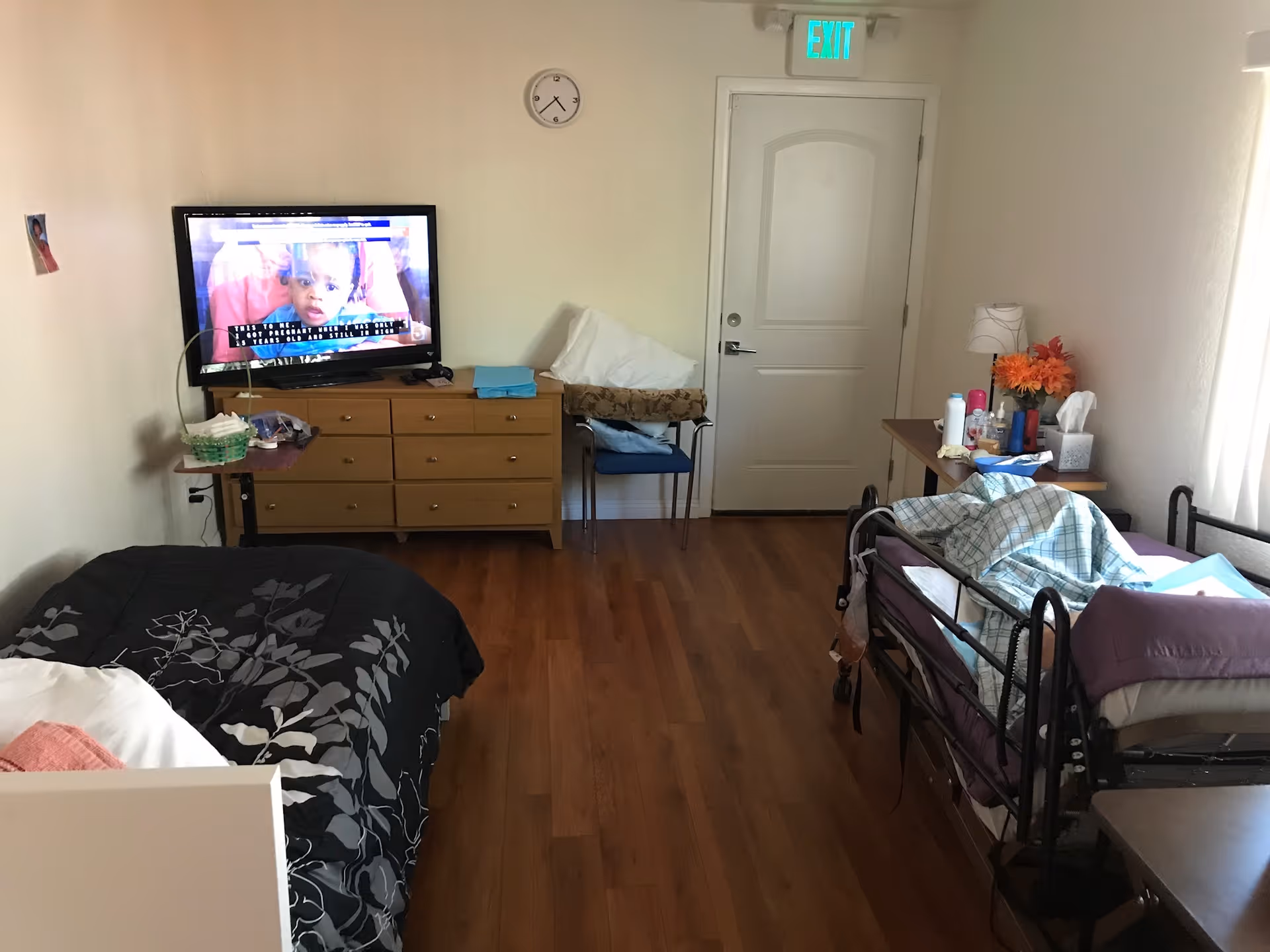 A room with two beds, one on the left with a black and gray floral blanket and one on the right with a metal frame and a blue checkered blanket. A wooden dresser with a TV on top is against the far wall, with a clock above it. There is a chair with folded linens and a table with various personal items and a lamp next to the bed on the right. The floor is wooden, and there is a white door with an exit sign above it at the back of the room.