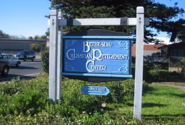 A blue wooden sign with white text that reads 'Bethesda Christian Retirement Center' mounted on white posts in a grassy area with bushes. There is a smaller blue sign below it that says 'Entrance 24/7'. Cars and buildings are visible in the background under a clear blue sky.