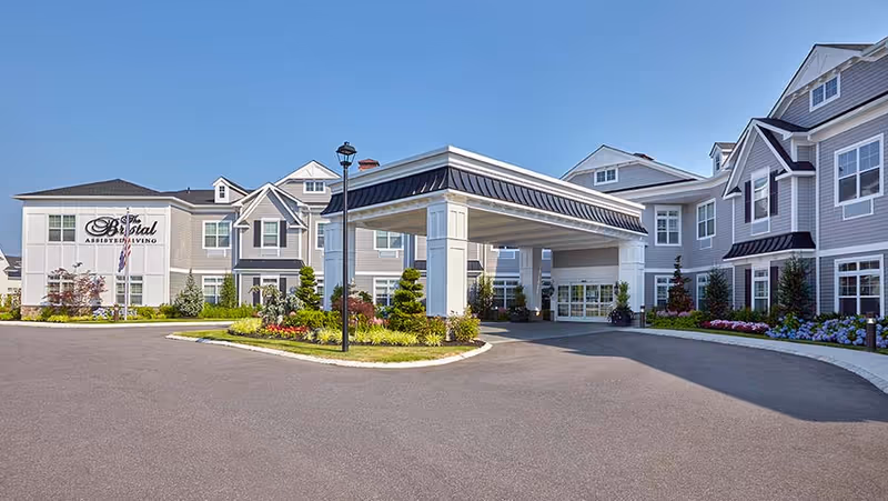 Exterior view of The Bristal Assisted Living at Mount Sinai building with a covered entrance, landscaped garden beds, and clear blue sky.