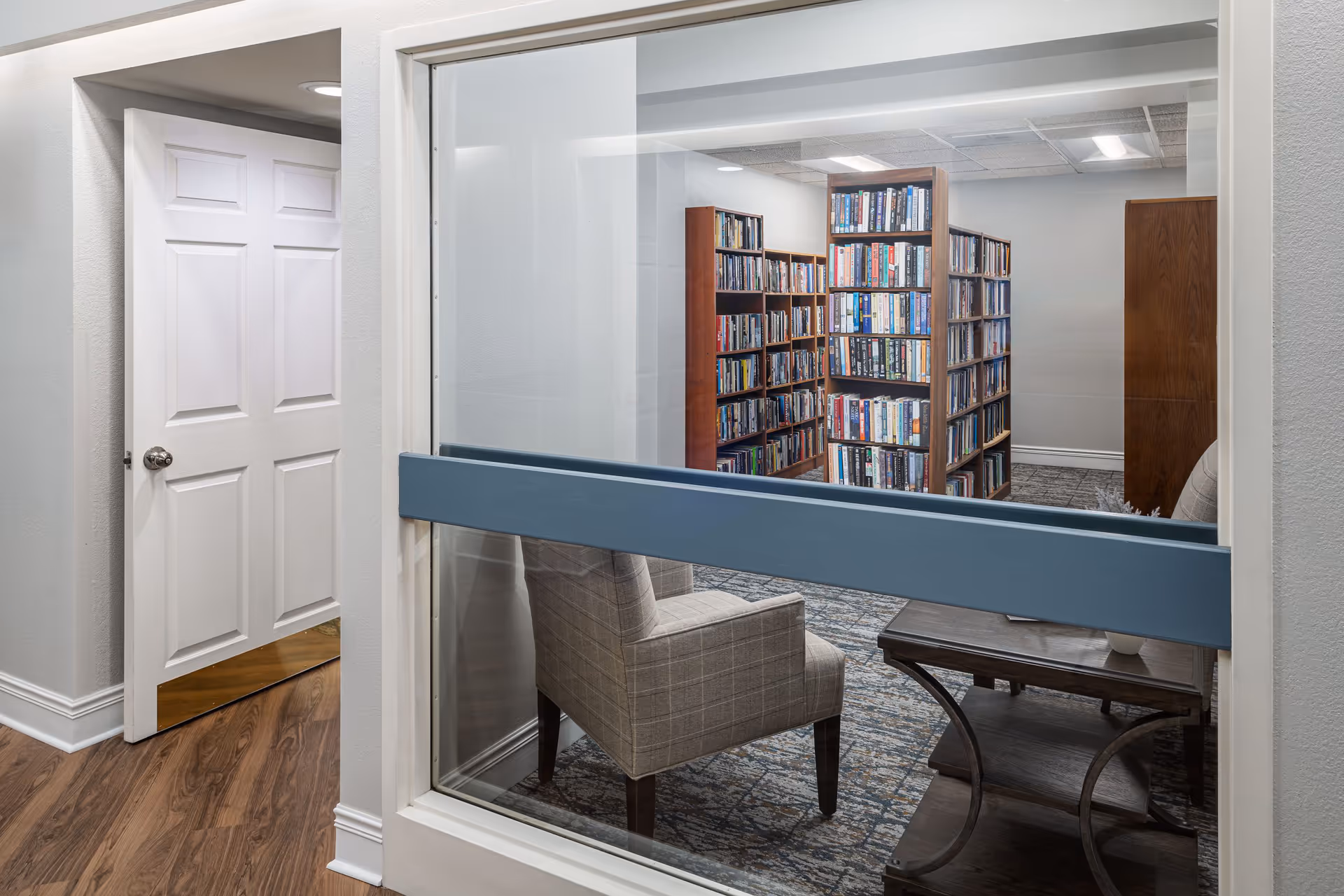 View through a large interior window into a quiet library room with bookshelves filled with books, a patterned carpet, and a seating area with a plaid armchair and a wooden side table. The hallway outside has wood flooring and a white door slightly ajar.