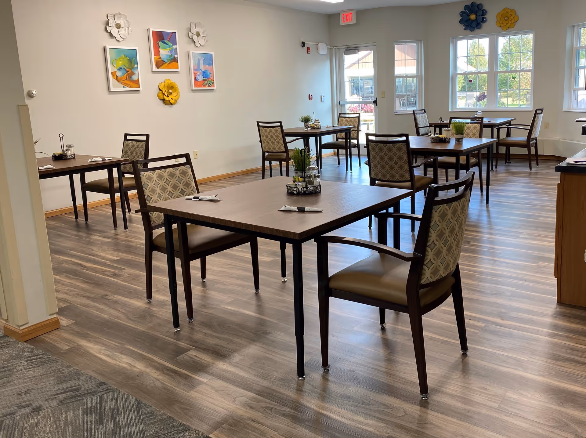 A bright dining room with several wooden tables and cushioned chairs arranged neatly. The room has large windows letting in natural light, colorful wall art, and decorative flowers on the walls. Each table has a small centerpiece and napkins with utensils.