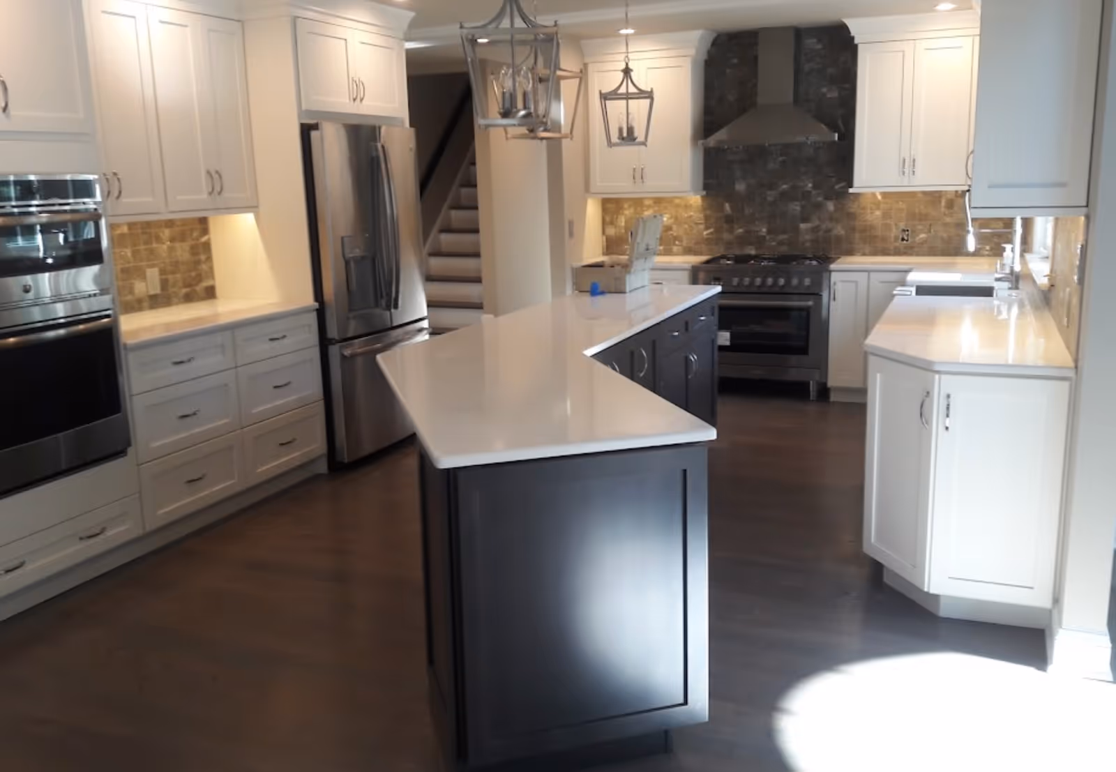 A modern kitchen with white cabinetry, stainless steel appliances including a refrigerator and oven, a large kitchen island with a white countertop, dark wood flooring, and pendant lights hanging above the island. There is a staircase visible in the background.