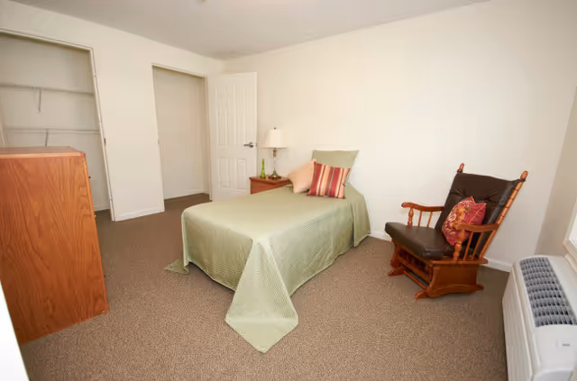 A simple bedroom with a single bed covered in a green bedspread and multiple pillows. There is a wooden rocking chair with a cushion, a wooden dresser, a nightstand with a lamp, an open closet, and a window air conditioning unit.