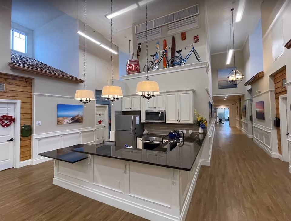 Interior view of a modern kitchen area in a senior living facility with white cabinets, stainless steel appliances, and a black countertop island with three hanging pendant lights. The space has high ceilings and wood flooring, with decorative items such as a bicycle, surfboards, and a Coca-Cola barrel displayed on a ledge above the cabinets. A hallway extends to the right with artwork on the walls and doors along the corridor.