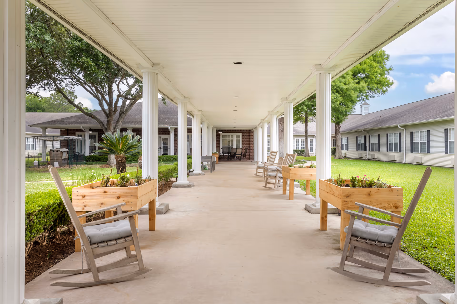Covered outdoor walkway with white columns and rocking chairs on both sides. Raised wooden planters with plants are placed near the chairs. The walkway is surrounded by green grass and trees, with buildings visible in the background under a partly cloudy sky.