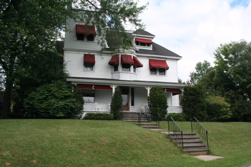 A large white multi-story house with red window awnings and a front porch, surrounded by green trees and bushes, with a grassy lawn and concrete steps leading up to the entrance.