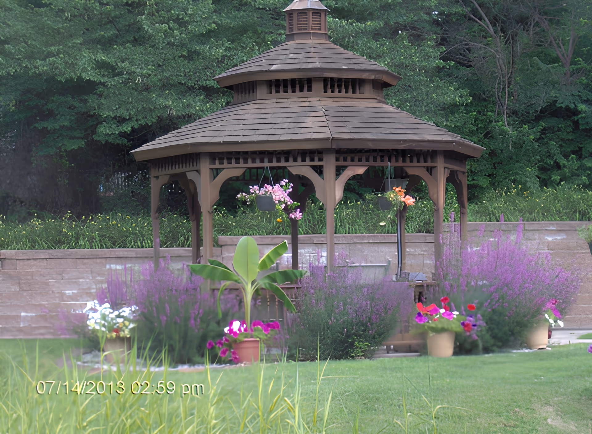 A wooden gazebo surrounded by various colorful flowers and plants in a garden area with a stone retaining wall and dense green trees in the background.