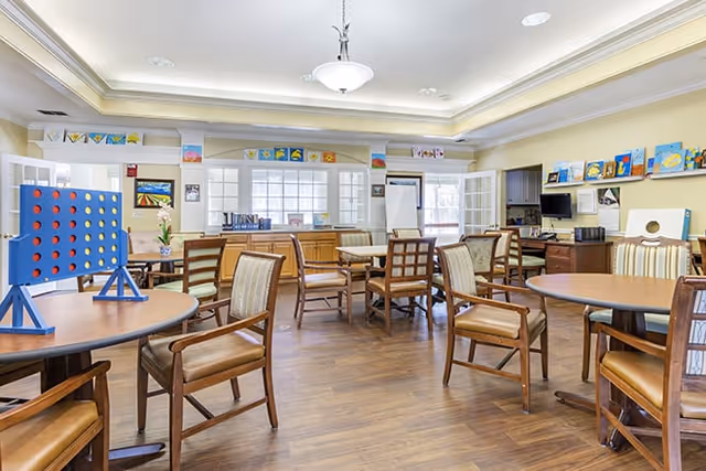 A bright and spacious common room with several round wooden tables and cushioned chairs arranged around the room. A large Connect Four game is set up on one table. The walls are decorated with colorful artwork and there are windows allowing natural light to fill the space. The room has a warm, inviting atmosphere with wood flooring and soft lighting.