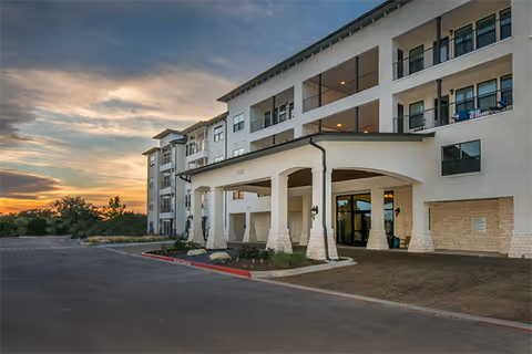 Exterior view of a multi-story senior living facility building at sunset with a covered entrance supported by stone pillars and a driveway in front.