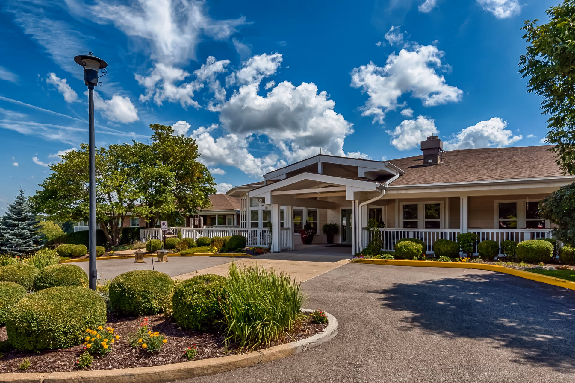 Exterior view of Autumn View Gardens Assisted Living and Memory Care facility showing the main entrance with a covered drop-off area, surrounded by well-maintained landscaping including bushes, flowers, and trees under a partly cloudy blue sky.
