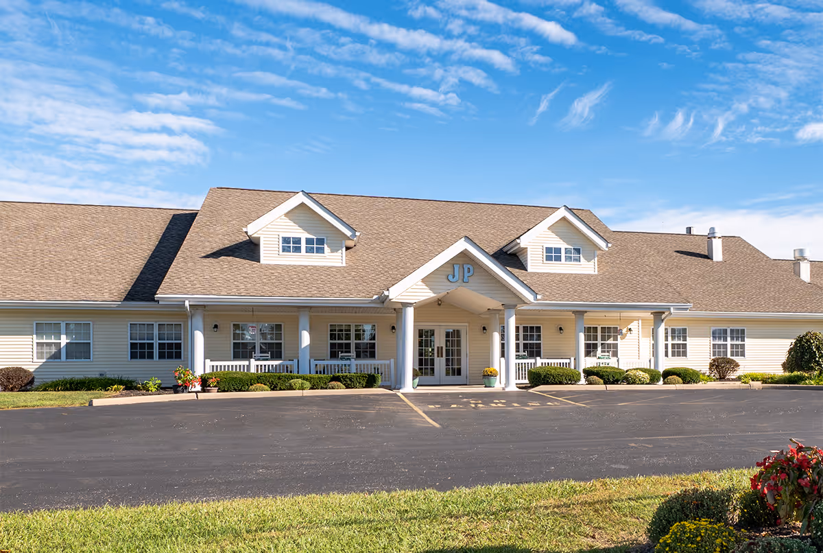 Front exterior view of a single-story senior living facility building with beige siding, a brown shingled roof, and a covered entrance supported by white columns. The building has multiple windows and small dormers on the roof. The letters 'JP' are displayed above the entrance. There is a paved parking area in front and some landscaping with bushes and flowers.