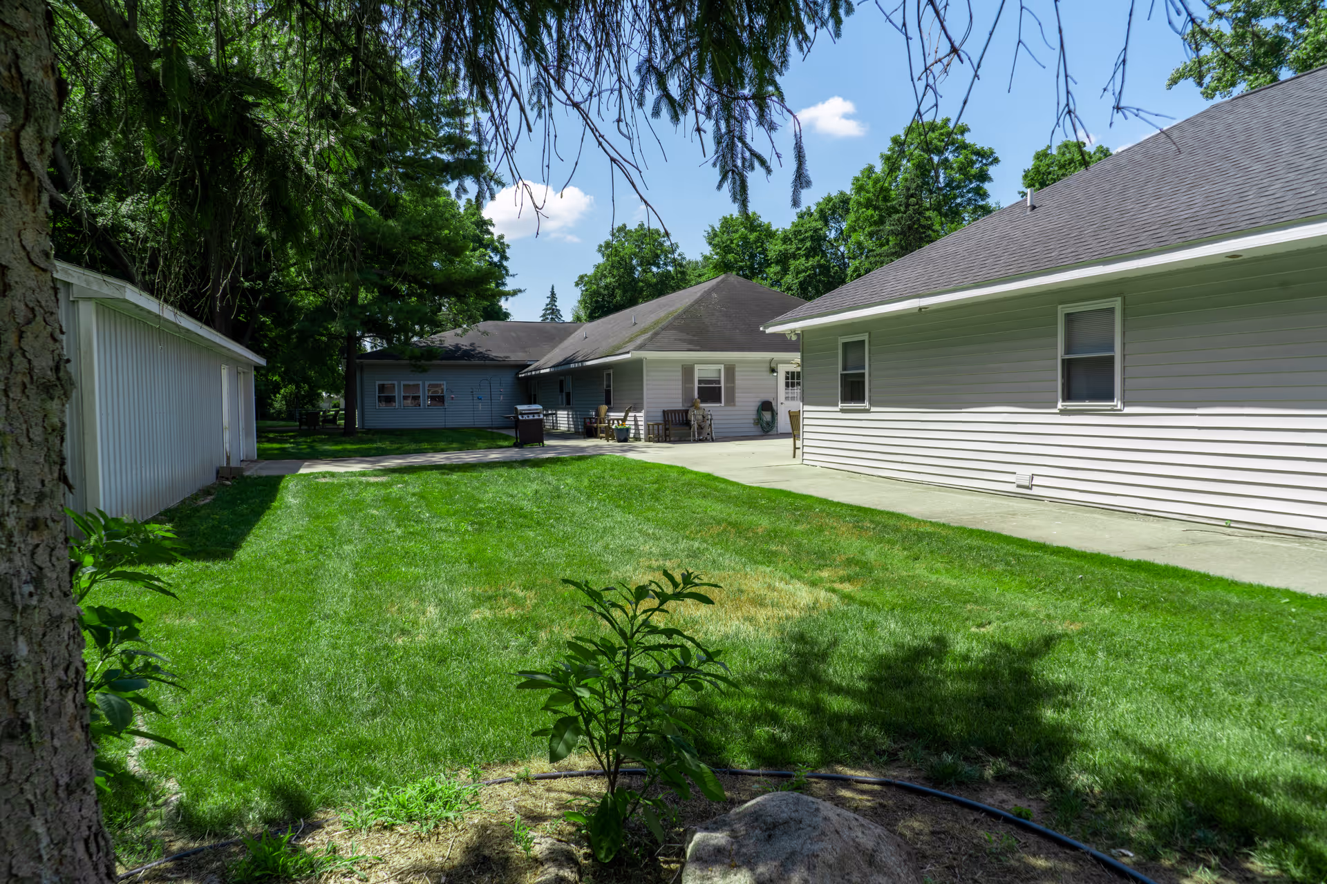 View of a green lawn area between two single-story buildings with light-colored siding under a blue sky with some clouds. Trees surround the area providing shade, and there is a concrete patio with outdoor furniture and a grill near the building in the background.