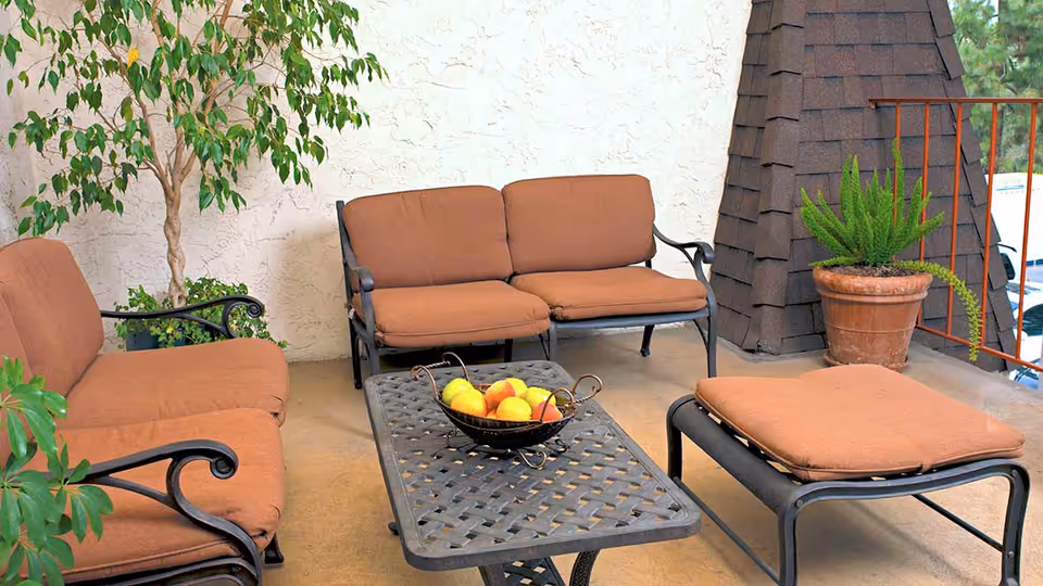 Outdoor patio area with brown cushioned metal seating including a loveseat, chair, and ottoman around a metal coffee table holding a bowl of fruit. There are potted plants and a textured white wall in the background.