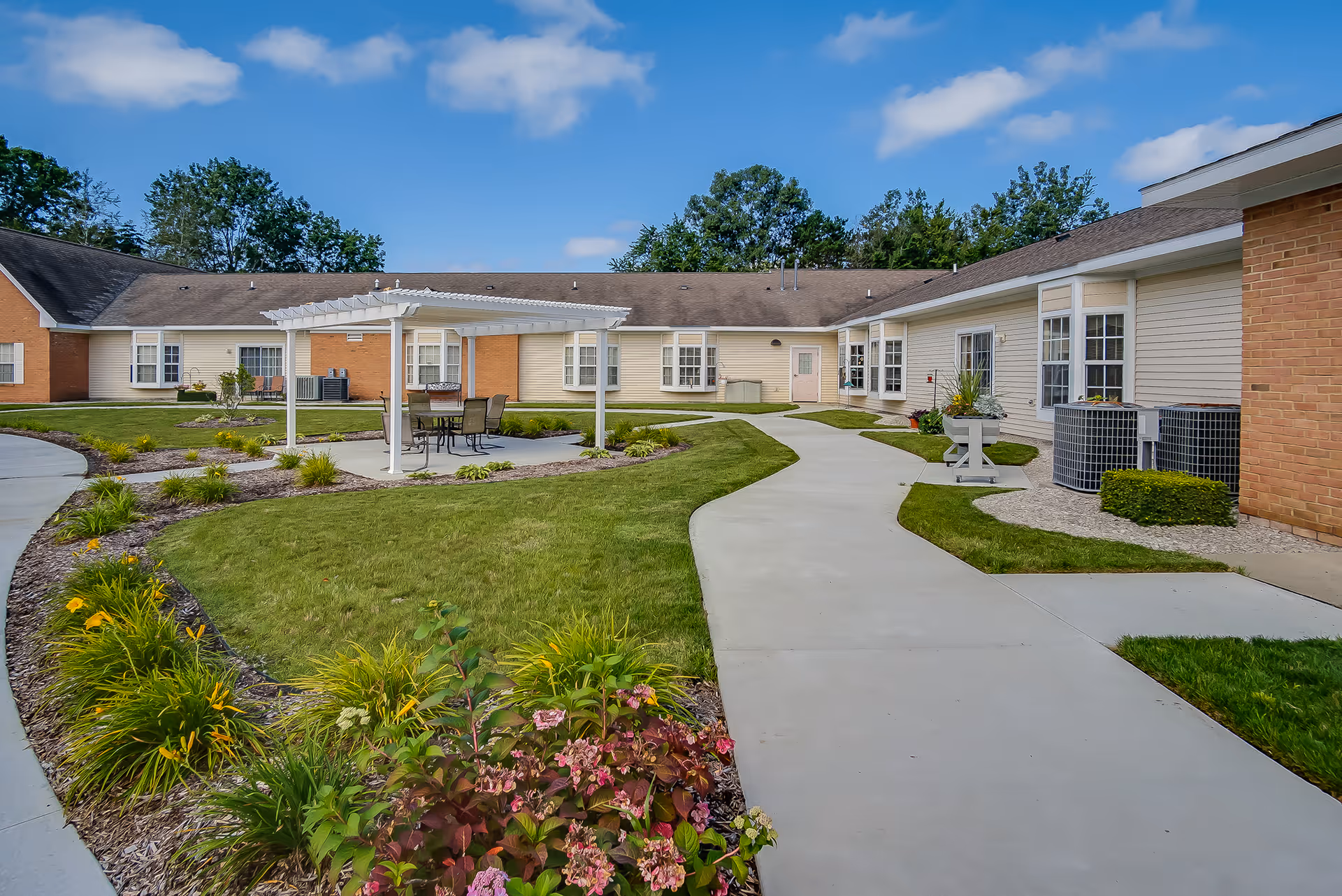 Outdoor courtyard area of River Ridge Retirement Village featuring a paved walkway, green lawn, flower beds, and a white pergola with a table and chairs underneath. The surrounding buildings have beige siding and brick accents under a blue sky with some clouds.