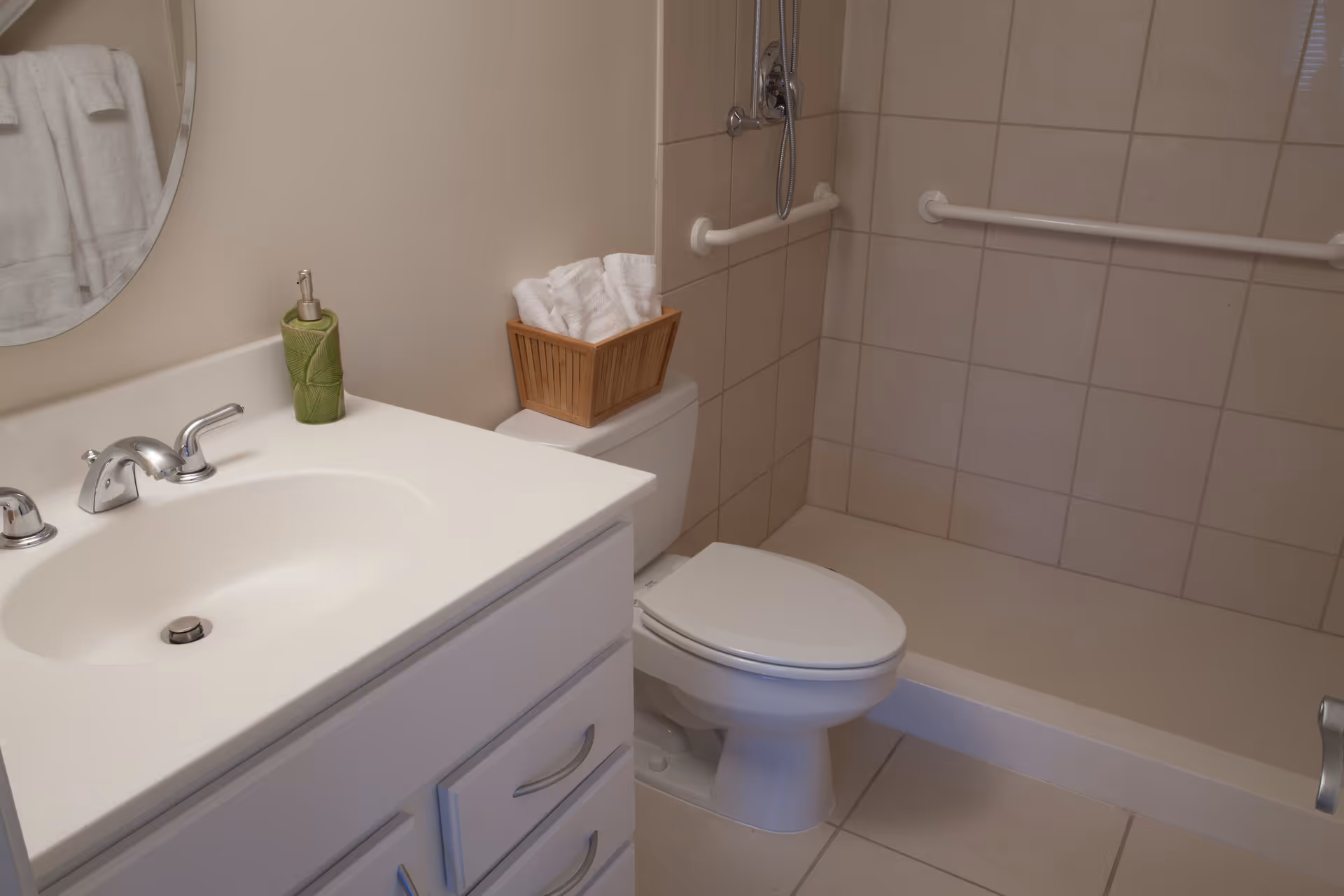 A clean bathroom featuring a white sink with a silver faucet, a green soap dispenser, a toilet with a basket of white towels on top, and a tiled shower area with grab bars installed on the walls.