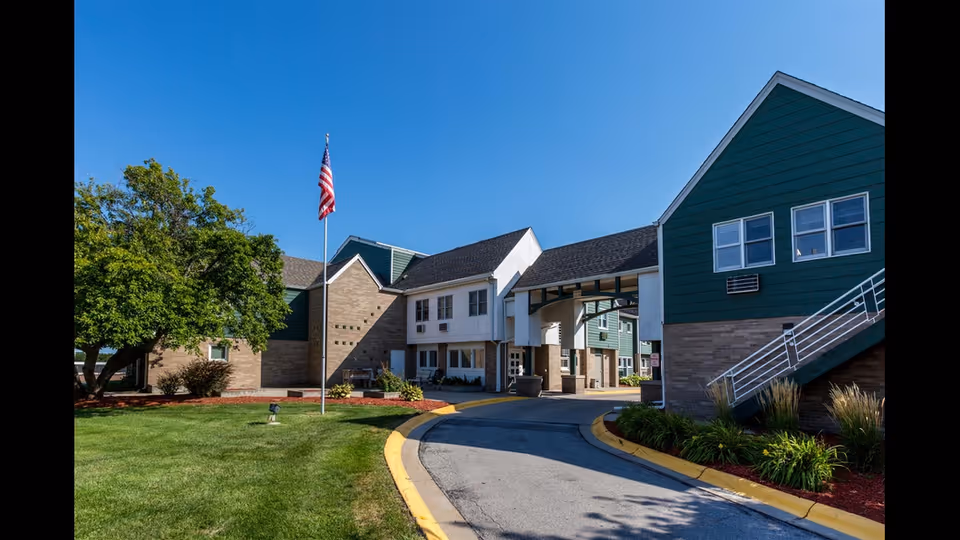 Exterior view of Hickory Villa Assisted Living facility showing a driveway leading to the entrance, green lawn with a tree, an American flag on a flagpole, and buildings with green and beige siding under a clear blue sky.