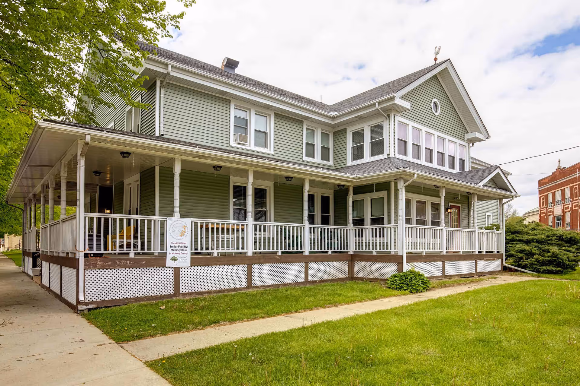 Exterior view of a two-story green senior living facility with white trim and a wraparound porch featuring white railings. The building is surrounded by a green lawn and a sidewalk, with trees and another building visible in the background under a partly cloudy sky.