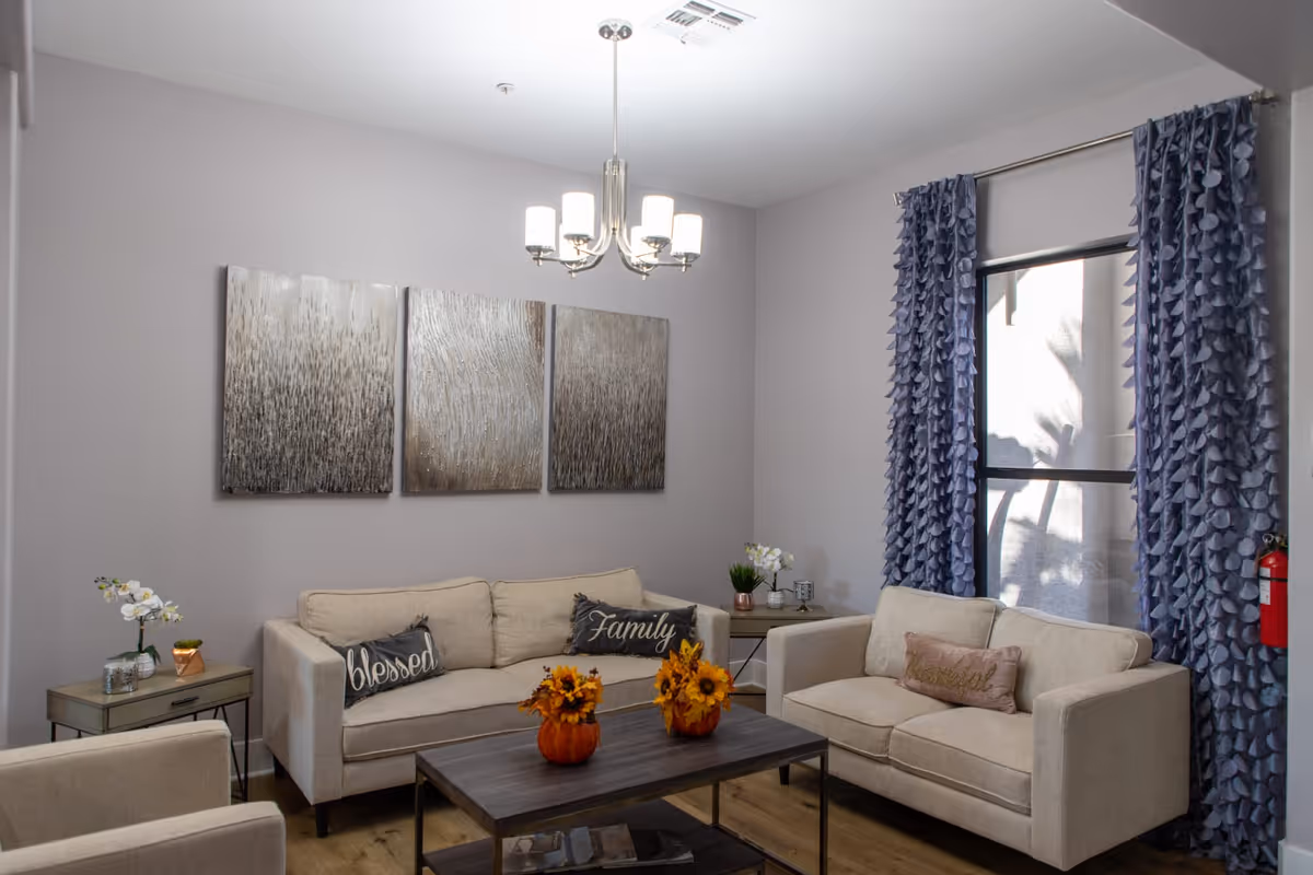 A cozy living room with beige sofas and armchairs, decorated with pillows that say 'blessed', 'Family', and 'thankful'. There is a wooden coffee table with two small pumpkin vases holding sunflowers. The walls are light gray with three abstract metallic art pieces hanging above the sofa. Blue ruffled curtains frame a window letting in natural light. Side tables hold small plants and decorative items, and a modern chandelier hangs from the ceiling.