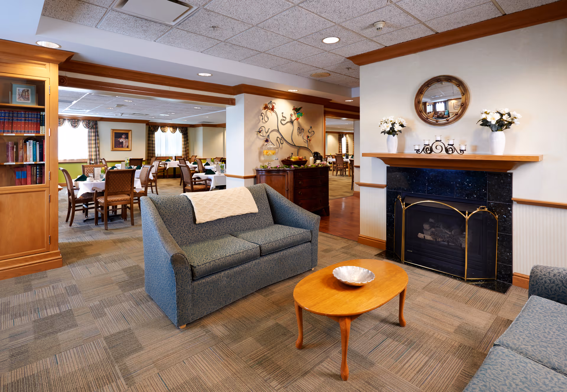 A cozy common area in a senior living facility featuring two blue upholstered sofas facing a wooden coffee table with a decorative bowl. There is a black marble fireplace with a wooden mantle adorned with two white vases of flowers, candles, and a round mirror above it. In the background, a dining area with multiple tables and chairs is visible, along with a wooden cabinet and decorative wall art.