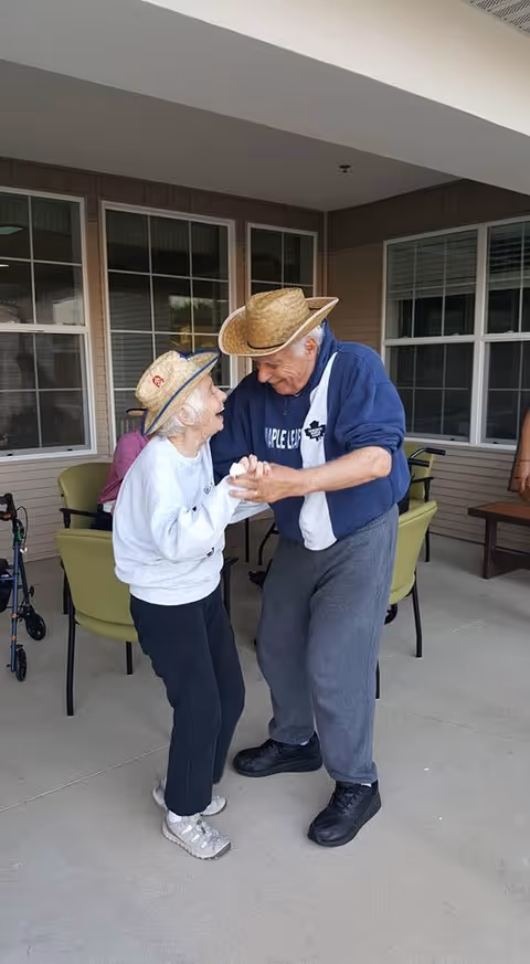 An elderly man and woman wearing straw hats are dancing together on a covered patio area outside a building. The woman is wearing a white sweatshirt, black pants, and sneakers, while the man is dressed in a blue jacket, gray sweatpants, and black shoes. Behind them are green chairs and windows of the building.
