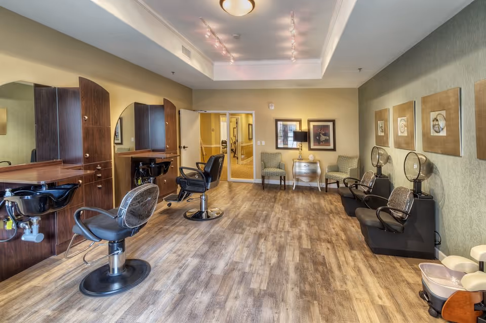Interior view of a salon area in a senior living facility with two styling chairs and wash basins on the left, two hair drying chairs on the right, and two armchairs with a small table and lamp against the far wall. The room has wood flooring, soft lighting, and framed artwork on the walls.
