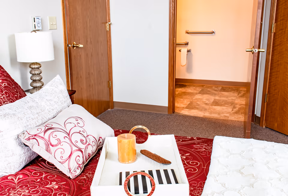 A bedroom with a bed covered in red and white patterned bedding and pillows. A white tray with a candle and a small decorative item is placed on the bed. A lamp with a white shade is on a nightstand next to the bed. Two wooden doors are visible, one leading to a bathroom with grab bars and a towel hanging on the wall.