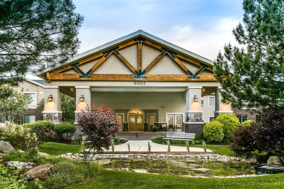 Front exterior view of a senior living facility with a covered entrance supported by stone and wood pillars, surrounded by well-maintained landscaping including trees, bushes, and a small pond with rocks in the foreground.