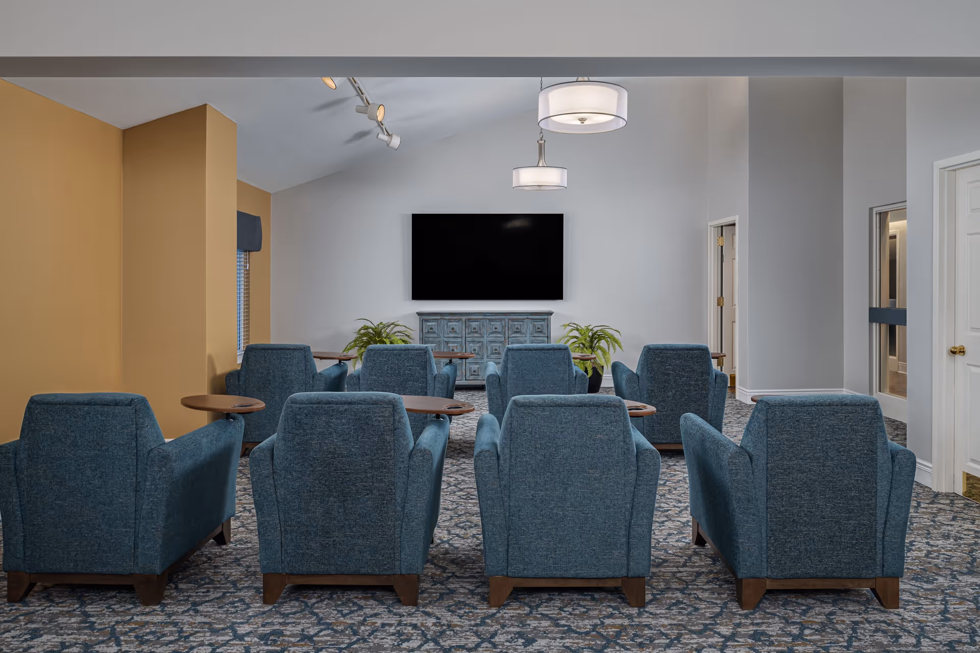A senior living facility common area with eight blue upholstered armchairs arranged in two rows facing a large flat-screen TV mounted on a white wall. Below the TV is a blue distressed cabinet with two potted plants on either side. The room has a patterned carpet, mustard yellow accent wall on the left, and two modern ceiling lights hanging from a vaulted ceiling.