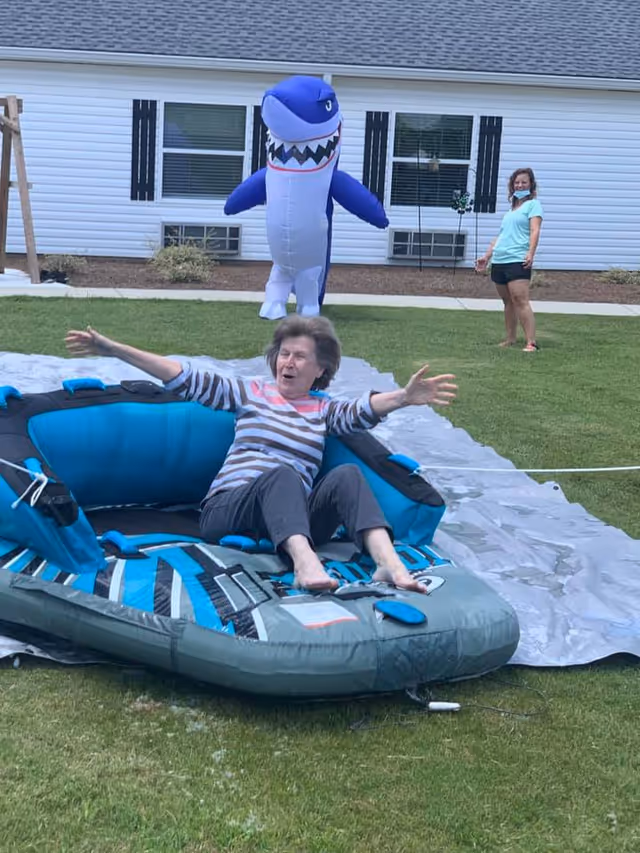 An elderly woman sitting on an inflatable water tube on a grassy lawn with her arms outstretched, appearing joyful. Behind her, a person wearing a face mask stands near a large inflatable shark figure. In the background is a white building with windows and black shutters.