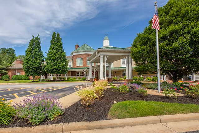 Exterior view of a senior living facility with a covered entrance, landscaped garden beds with flowers and shrubs, an American flag on a flagpole, and a clear blue sky with some clouds.