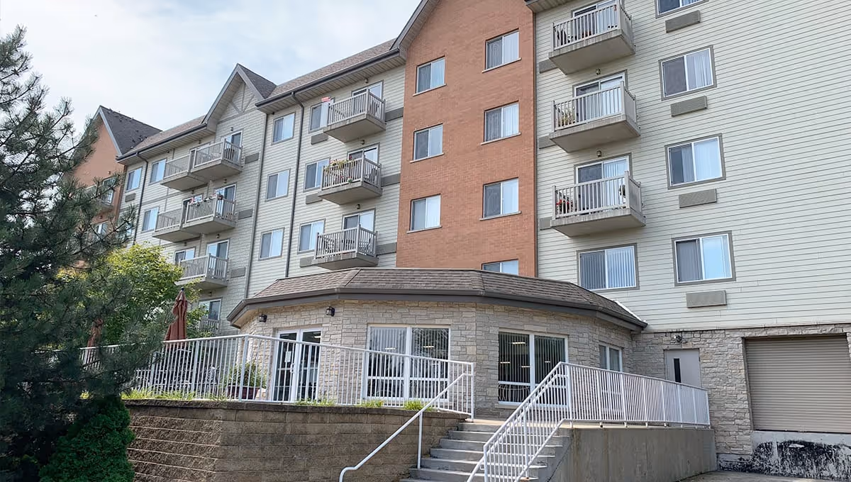 Exterior view of a multi-story senior living facility with balconies, a stone and siding facade, and a staircase leading to a patio area surrounded by a white railing. Trees and greenery are visible around the building.