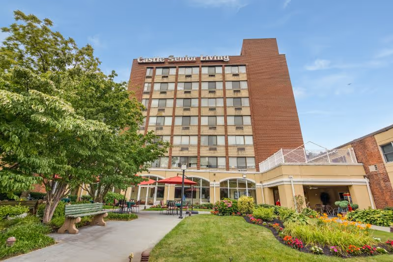 Exterior view of Castle Senior Living, a multi-story brick building with several windows. In front of the building is a well-maintained garden with green grass, colorful flowers, trees, benches, and outdoor tables with red umbrellas under a clear blue sky.