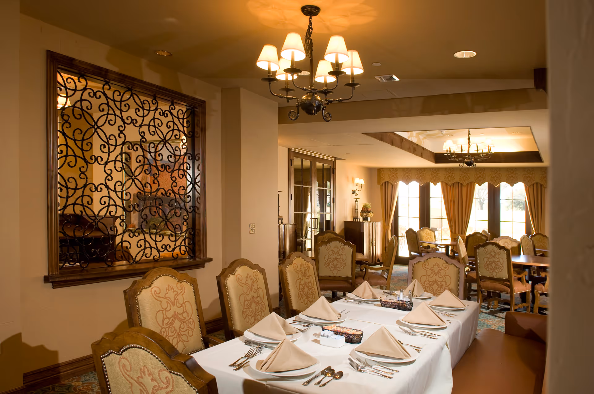 A warmly lit dining room with a long table set for eight people. The table is covered with a white tablecloth and neatly arranged with beige folded napkins, plates, and silverware. Ornate wooden chairs with patterned upholstery surround the table. In the background, there are more tables and chairs, large windows with curtains, and decorative wall elements including a wrought iron framed window and a chandelier hanging from the ceiling.