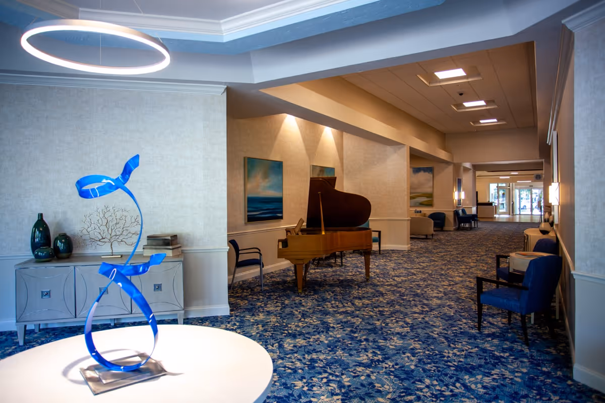 Interior view of a senior living facility hallway with blue patterned carpet, a grand piano, several chairs, and wall art. A modern blue sculpture is displayed on a round white table in the foreground, and decorative vases and books are on a sideboard against the wall.