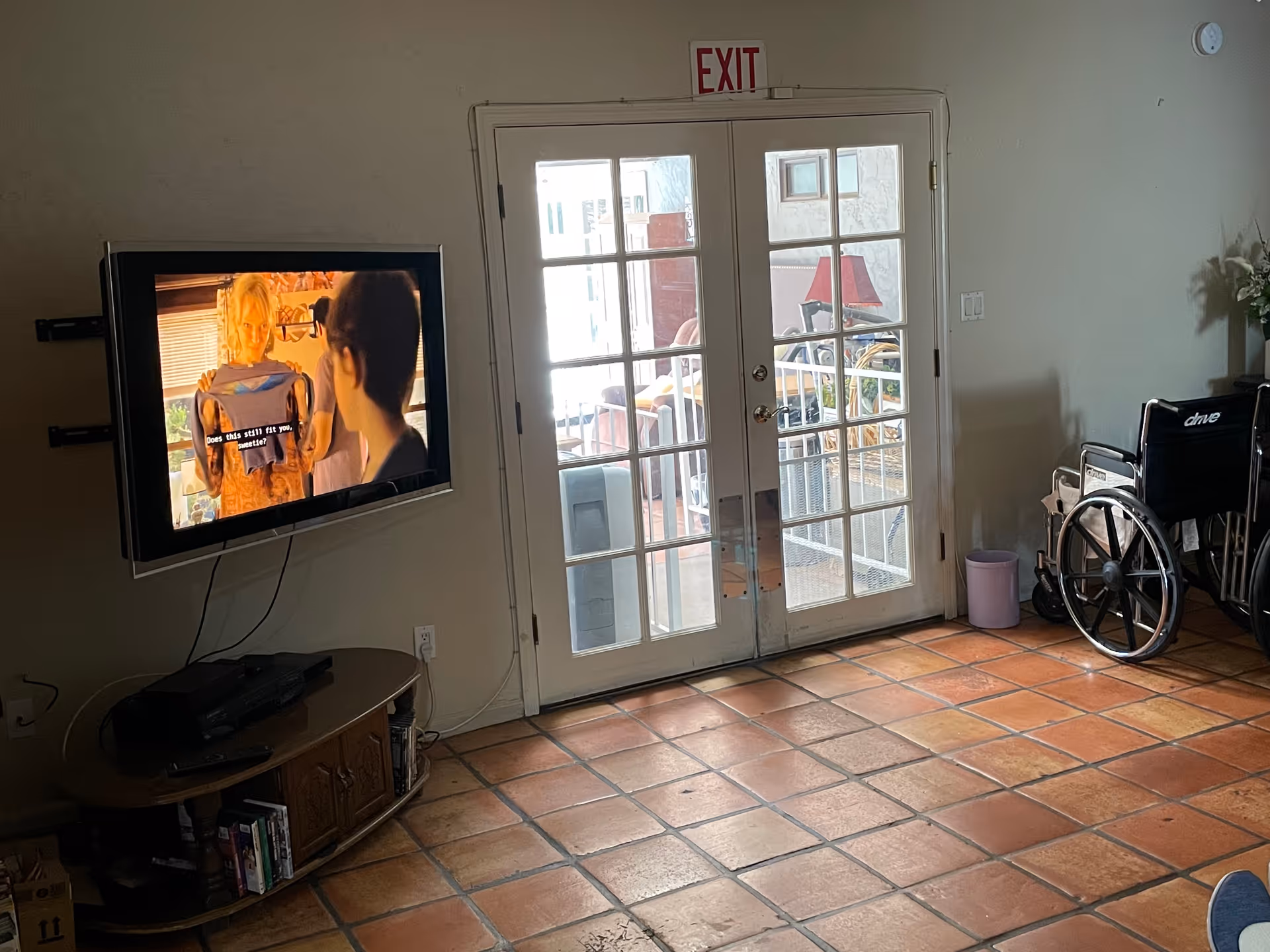 Interior room with terracotta tiled floor, a wall-mounted TV showing a scene with subtitles, a wooden cabinet below the TV, a wheelchair near a set of glass double doors with an exit sign above, and some furniture visible outside through the doors.