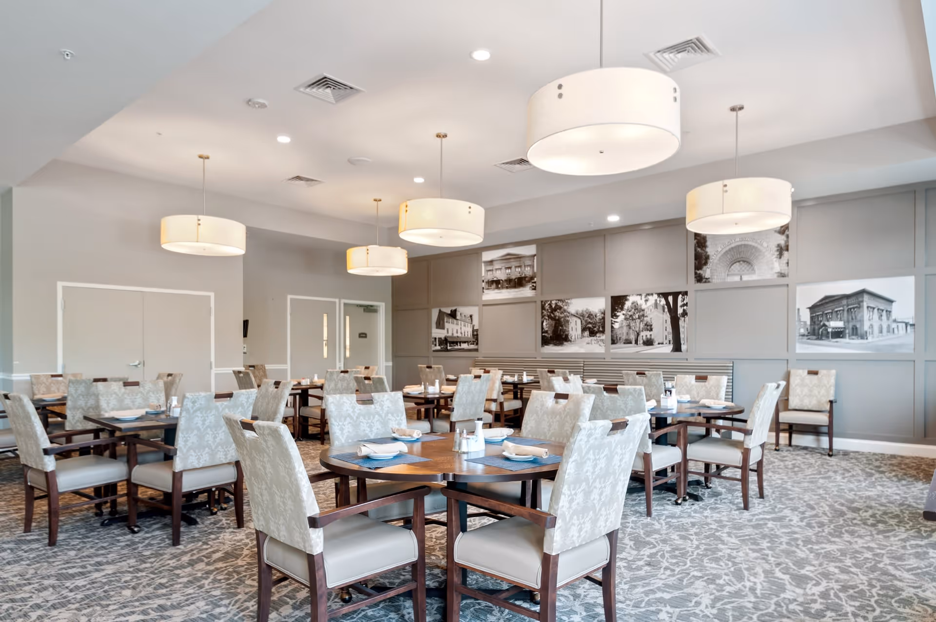 A spacious dining room with multiple round wooden tables and cushioned chairs arranged neatly. The room features modern hanging light fixtures, a patterned carpet, and black and white framed photographs on the walls.