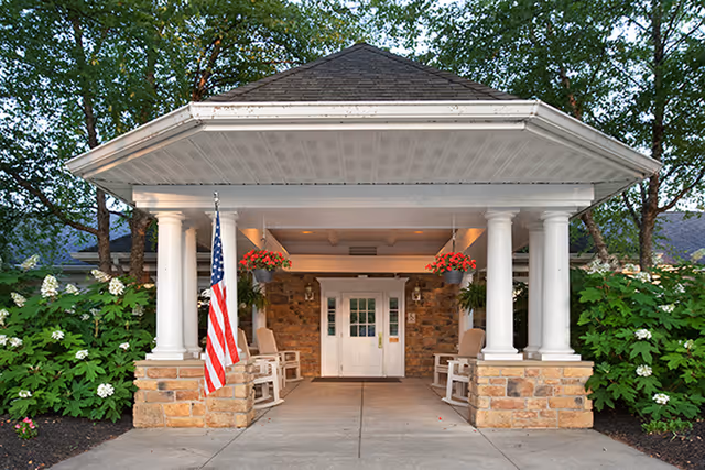 Entrance of a building with a covered porch supported by white columns and stone bases, featuring an American flag on the left side, hanging flower baskets, rocking chairs, and a white double door under the porch roof. Green bushes and trees surround the entrance.